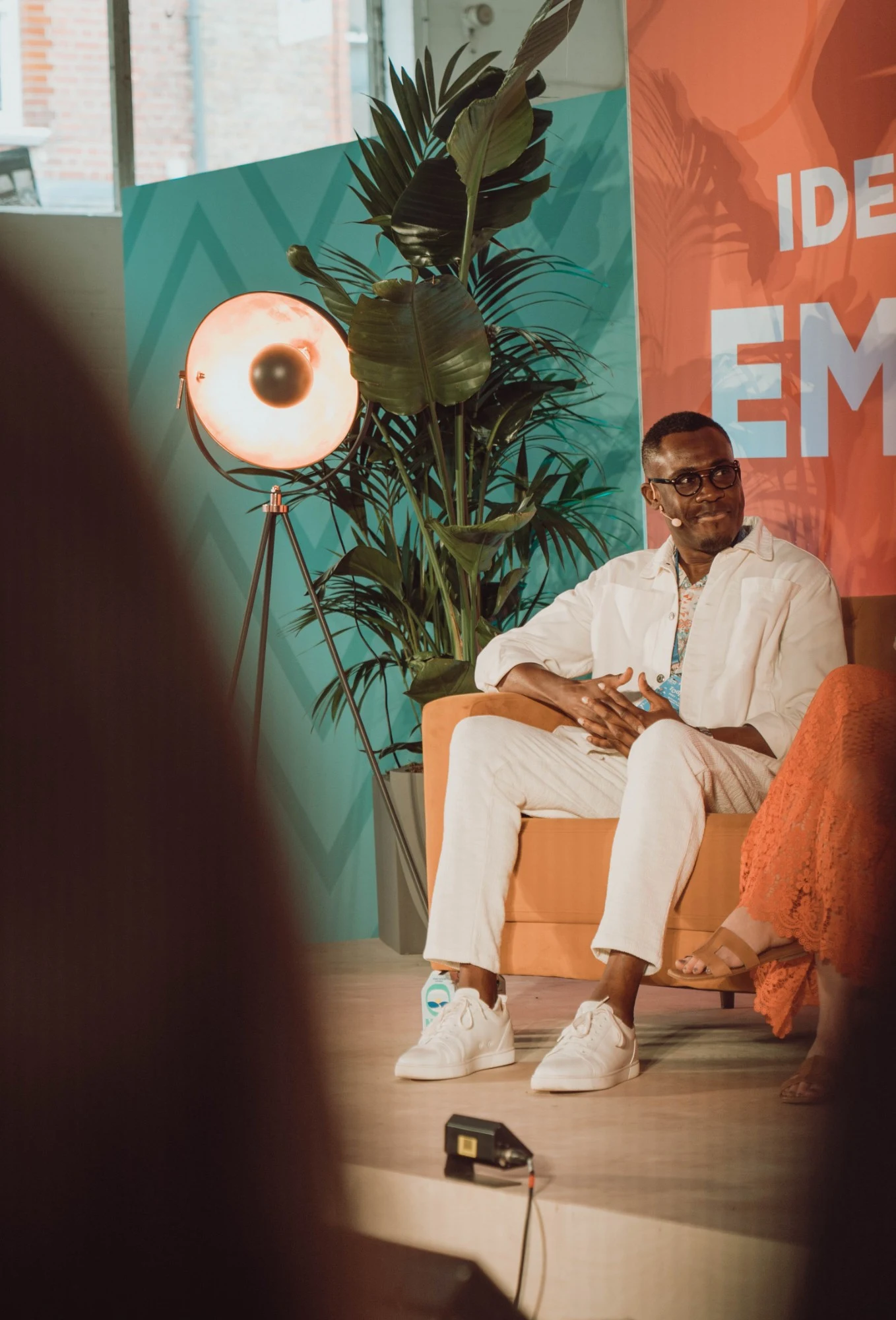 Peter Komolafe smiling on an orange sofa on stage, a studio lamp and plants beside him