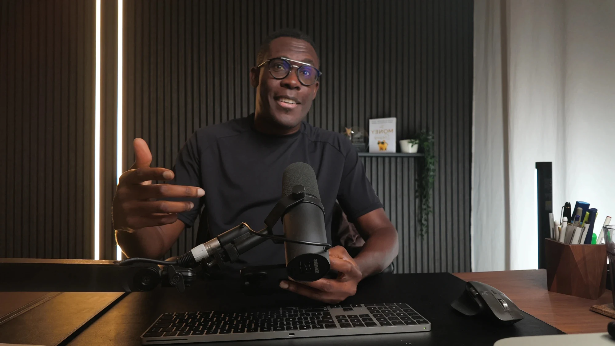Peter Komolafe at his desk in the studio, mid-sentence behind his podcast microphone, his book on the shelf behind