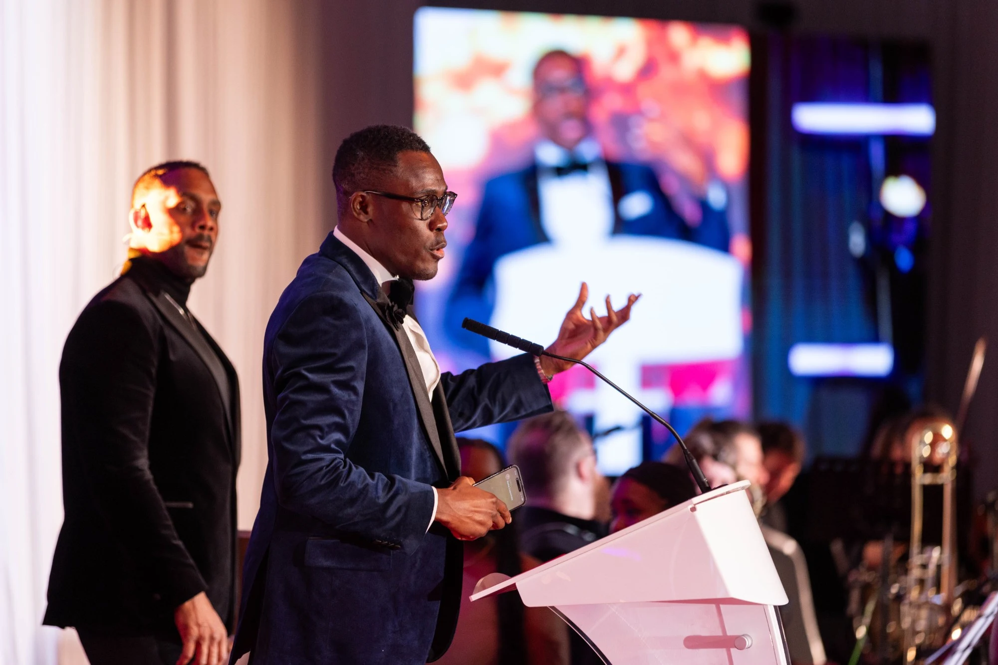 Peter Komolafe speaking at a podium on stage at an awards evening, a band behind him